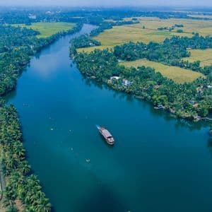 Una veduta aerea di una casa galleggiante che naviga lungo un ampio fiume azzurro, circondata da rigogliose palme verdi e campi agricoli.