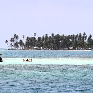 Un groupe WeRoad se baigne dans les eaux peu profondes et turquoise d'un banc de sable, avec leur bateau amarré à proximité et une île couverte de palmiers en arrière-plan.