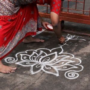 Une femme en sari rouge à motifs dessine un grand motif floral blanc sur le trottoir avec de la poudre.