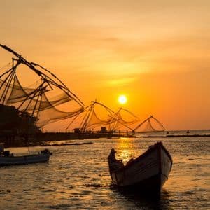 Ein kleines Boot mit einer Person liegt auf dem Wasser nahe chinesischer Fischernetze, die sich vor einem goldenen Sonnenuntergang abzeichnen.