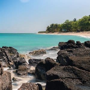 Lunga esposizione di acqua turchese che scorre su rocce scure su una spiaggia tropicale sotto un cielo azzurro chiaro.
