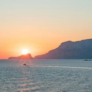 Una piccola barca naviga sul mare mentre il sole tramonta dietro un'isola in silhouette, proiettando un bagliore arancione sull'acqua.