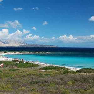Una vista panoramica di una spiaggia di sabbia bianca e mare turchese, con una grande isola sullo sfondo sotto un cielo parzialmente nuvoloso.