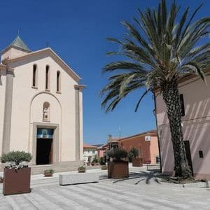 Una chiesa rosa pallido e una grande palma si ergono in una piazza cittadina soleggiata e pavimentata sotto un cielo azzurro e limpido.