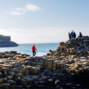 Un voyage de groupe WeRoad explore une formation côtière de colonnes de basalte entrelacées, au bord de la mer, sous un ciel ensoleillé.