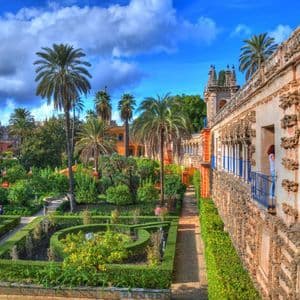 Une vue plongeante sur un jardin à la française luxuriant avec des palmiers et des haies sculptées, à côté d'un bâtiment en pierre orné sous un ciel partiellement nuageux.