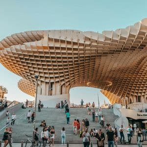 Des personnes marchent et se rassemblent sur de larges escaliers sous un grand auvent architectural moderne en treillis, sous un ciel dégagé.