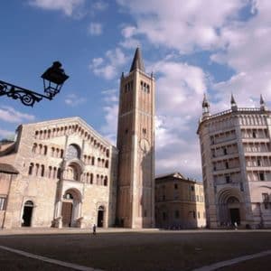 Una cattedrale storica, un campanile e un battistero si ergono in un'ampia piazza pubblica sotto un cielo parzialmente nuvoloso.