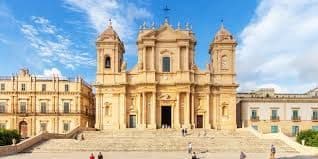 La facciata ornata, color sabbia, di una cattedrale barocca in cima a un'ampia scalinata sotto un cielo blu con nuvole sottili.