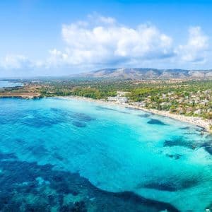Vista aerea di un mare turchese che incontra una spiaggia sabbiosa con una cittadina costiera, colline verdi sullo sfondo e un cielo azzurro.