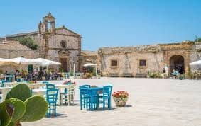 Un caffè all'aperto con tavoli e sedie di un blu brillante in una soleggiata piazza in pietra, di fronte a edifici storici.