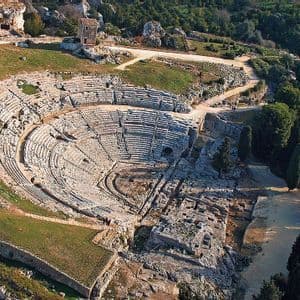 Veduta aerea di un antico anfiteatro in stile romano scavato in una collina rocciosa e circondato da alberi verdi.