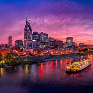 An illuminated riverboat floats on a wide river in front of a modern city skyline under a vibrant purple and pink sky at twilight.
