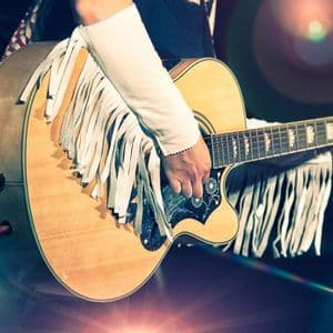 A close-up of a person wearing a white fringed sleeve playing an acoustic guitar on a stage with lens flares.