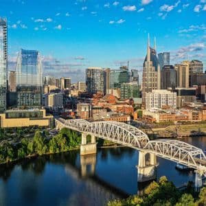 An aerial view of a modern city skyline with skyscrapers and a white truss bridge crossing a river under a blue sky.