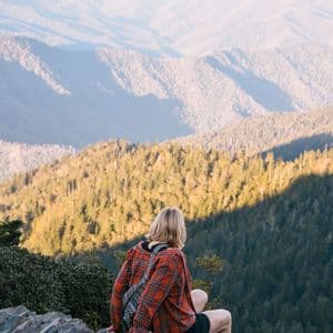 A person in a red plaid shirt sits on a rocky ledge, looking out over a vast mountain range covered in trees.