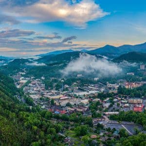 A panoramic aerial view of a town nestled in a green mountain valley with low clouds and mist under a blue sky.