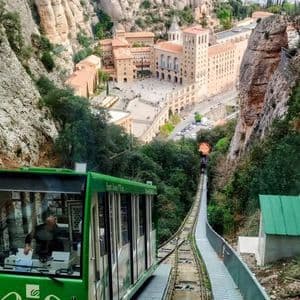 A high-angle view from a green funicular descending a steep track towards a large monastery complex built into a rocky mountain.