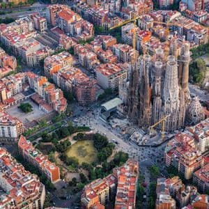 An aerial view of a large, ornate cathedral under construction, surrounded by a dense grid of city buildings with red roofs.