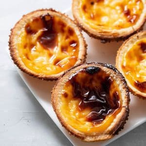 A close-up of several Portuguese custard tarts, pastéis de nata, with caramelized tops on a white rectangular plate.