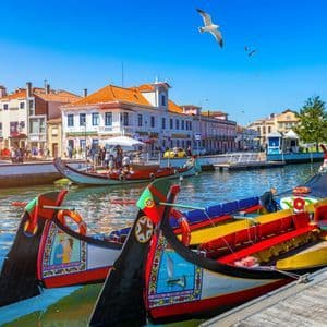 Colorful traditional boats are moored in a city canal, with waterfront buildings and people in the background under a clear blue sky.