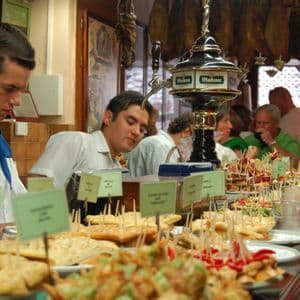A long wooden counter in a bustling bar, laden with a wide variety of tapas and pintxos for customers.
