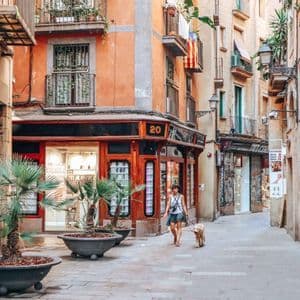 A woman walks her dog on a leash down a narrow cobblestone alley flanked by old buildings with balconies.