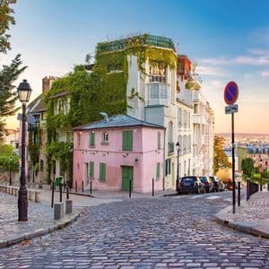A pink house with green shutters sits on a cobblestone street corner next to an ivy-covered building at sunset.