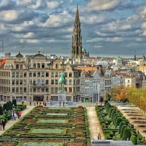 An elevated view of a formal garden with manicured hedges and pathways, leading towards a historic cityscape with a tall spire under a cloudy sky.
