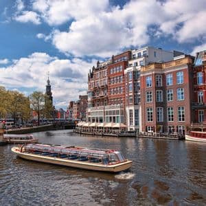 A tour boat with a glass roof cruises down a city canal lined with traditional brick buildings under a partly cloudy sky.