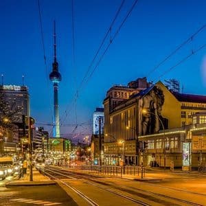 A city street at night with illuminated buildings, tram tracks, and a tall television tower against a dark blue sky.