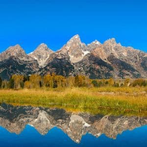 A jagged mountain range with patches of snow is reflected perfectly in a calm lake, with autumn foliage on the shore.