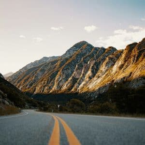 A low-angle shot of a winding asphalt road with yellow lines running through a mountain valley at golden hour.