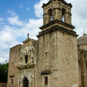 A weathered stone mission church with an ornate facade and bell tower under a partly cloudy sky.