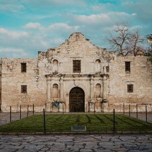 The facade of a historic stone mission building standing behind a cobblestone plaza under a cloudy blue sky.