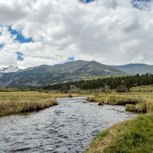 A wide river winds through a green meadow towards distant, snow-capped mountains under a partly cloudy sky.