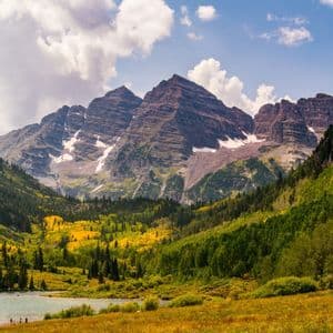 A mountain range with snow patches overlooks a valley filled with autumn-colored forests and a lake, under a partly cloudy sky.