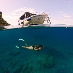 Una vista sopra e sotto il livello dell'acqua mostra una persona fare snorkeling in acque cristalline sotto una piccola imbarcazione ormeggiata in mare.