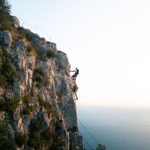 Un viaggio di gruppo WeRoad con due persone, caschi e imbracature, che praticano arrampicata su roccia su una parete a strapiombo sul mare.