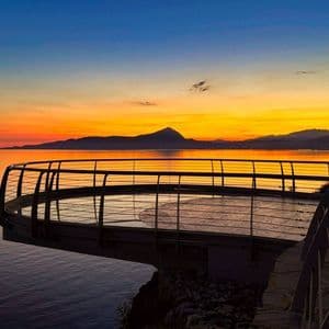Una piattaforma di osservazione circolare si estende su un mare calmo al tramonto, con montagne lontane stagliate contro il cielo.
