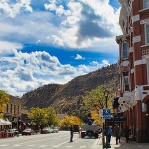 A wide street in a historic town with brick buildings and autumn trees, with a mountain rising in the background under a cloudy blue sky.