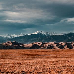 A dry, grassy plain leads to rolling sand dunes with a range of snow-capped mountains in the background under a cloudy sky.