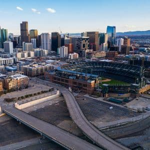 An aerial view of a large baseball stadium and empty roads with a modern city skyline and mountains in the background.