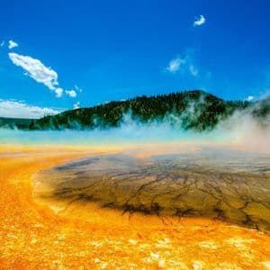 Vibrant orange mineral deposits surround a steaming geothermal hot spring with a forested hill under a bright blue sky.