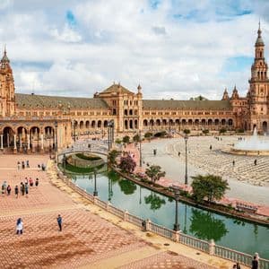 Vue d'en haut d'une grande place avec un édifice semi-circulaire orné, un canal, une fontaine et des passants.