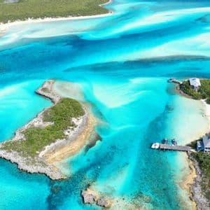An aerial view of small, green islands and sandbars surrounded by vibrant turquoise water, with a few houses and a pier.
