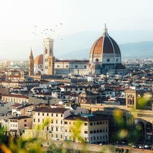 Un paesaggio urbano illuminato dal sole, con al centro una grande cattedrale dalla cupola in terracotta e un campanile, mentre uccelli volano nel cielo.