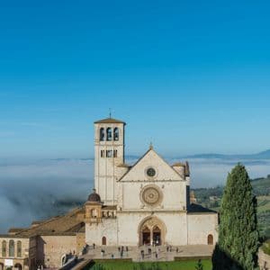 Una grande basilica in pietra con un campanile si affaccia su una valle ricoperta di nuvole basse sotto un cielo azzurro e limpido.