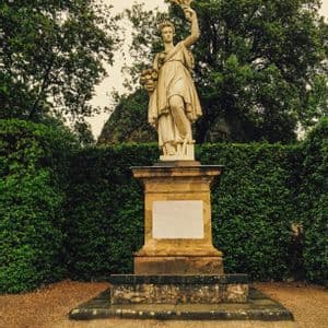 Una statua classica in pietra di una donna che tiene della frutta si erge su un piedistallo in un giardino con siepi verdi.