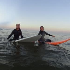Due donne del gruppo WeRoad, in mute, sorridono sedute sulle loro tavole da surf nell'oceano.